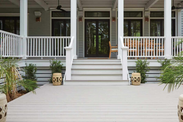 A spacious and clean white deck leading up to a home's entrance with white railing and stairs. The deck features two decorative stone planters, lush green plants, and a rocking chair visible through the glass door, complemented by the home's grey siding and white trim.
