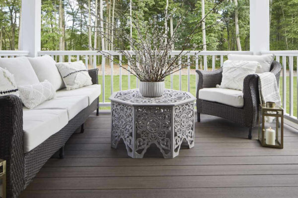 A cozy outdoor seating area on a dark wooden deck featuring a white cushioned sofa and chair with woven frames, white decorative pillows, an ornate white metal coffee table with a vase of pussy willow branches, all overlooking a forested backyard, with a white railing and a lantern on the side.